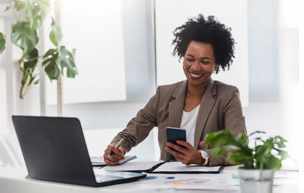 Zakelijke vrouw glimlacht terwijl ze op haar smartphone kijkt aan een bureau met laptop, documenten en planten in een moderne kantooromgeving.
