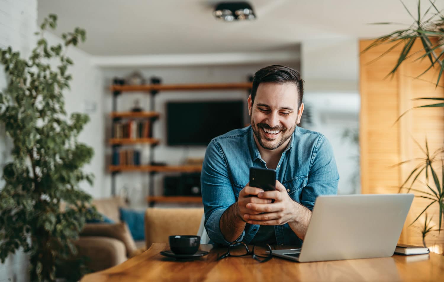 Glimlachende man werkt vanuit huis aan een houten tafel met laptop en smartphone, in een sfeervolle woonkamer met planten en boekenplank.