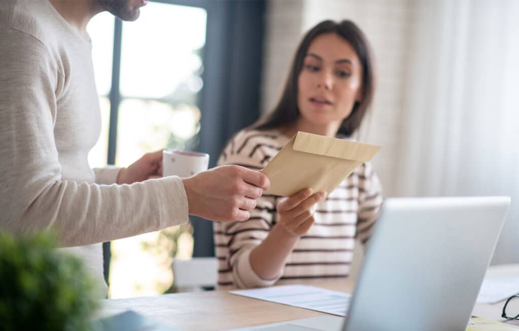 Twee collega’s hebben post ontvanger op kantoor; een vrouw bekijkt een bruine envelop terwijl een man met een mok erbij staat met een laptop op tafel.