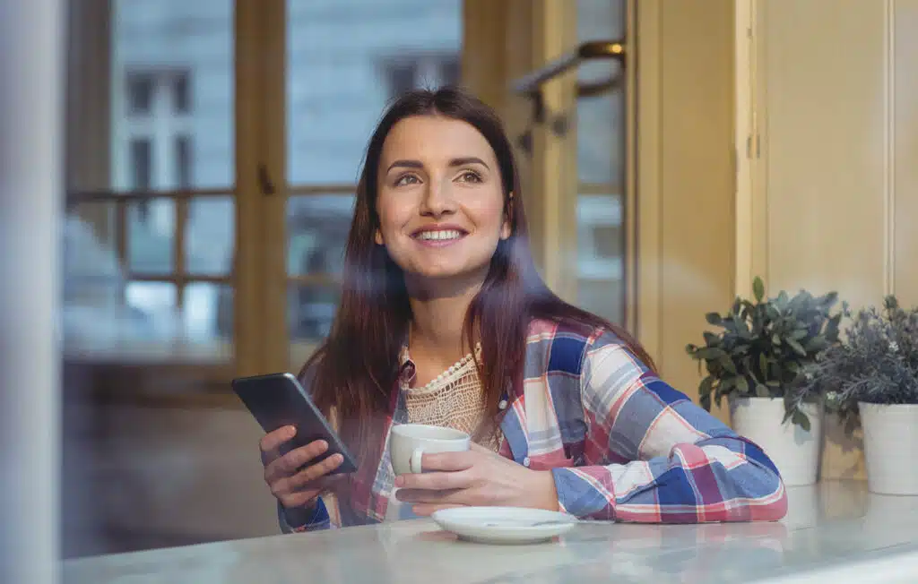 Vrouw die naar buiten kijkt met haar telefoon in de hand en een kop koffie in de andere.