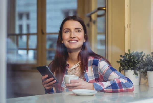 Vrouw die naar buiten kijkt met haar telefoon in de hand en een kop koffie in de andere.