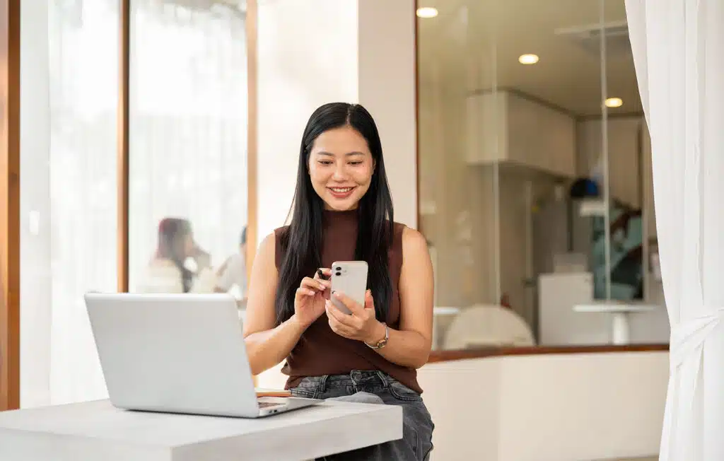 A satisfied Asian woman checks messages on her smartphone while working remotely from a coffee shop.